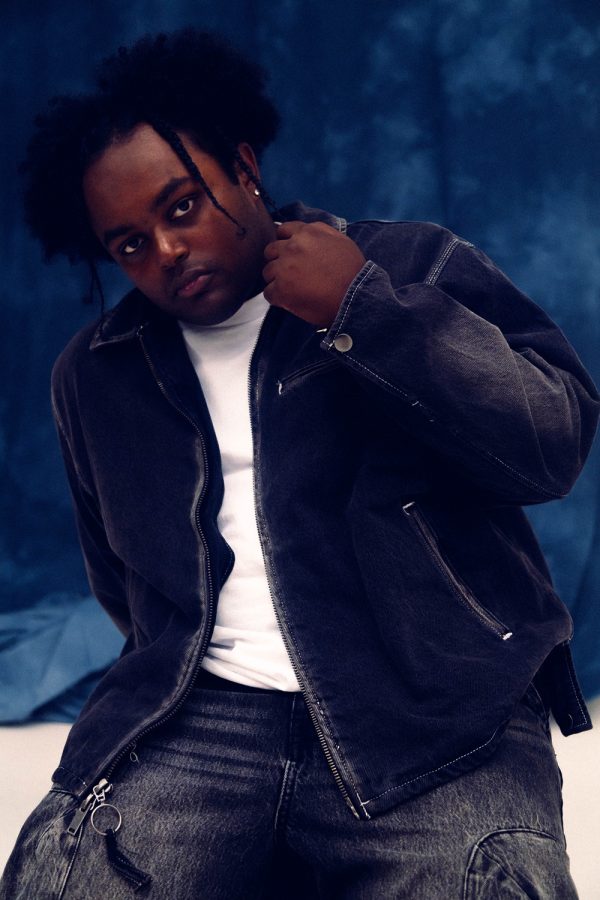 Youthful Black male model wearing a black denim jacket and white t-shirt, posing confidently against a dark blue background, showcasing modern street fashion and urban style for magazine or fashion brand imagery.