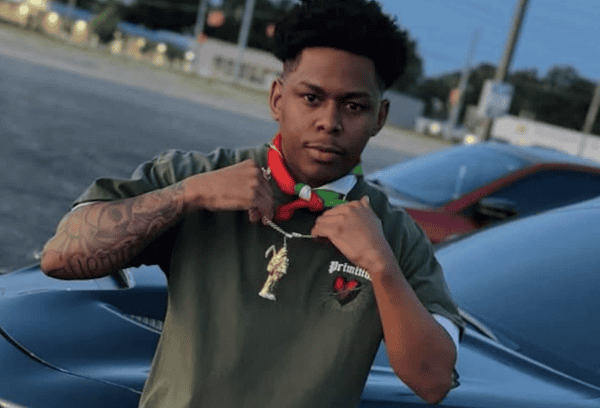 Young man showing off jewelry near his neck and arm, standing outdoors during twilight with street and car in the background, wearing a dark t-shirt with nostalgic patches.