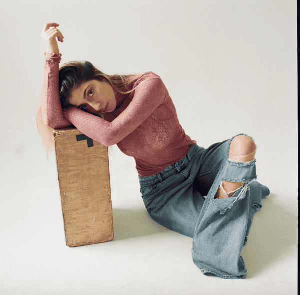 Young woman with long hair in trendy ripped jeans and pink lace top, sitting on the floor with her head resting on a wooden block, looking contemplative in a minimalist studio setting.