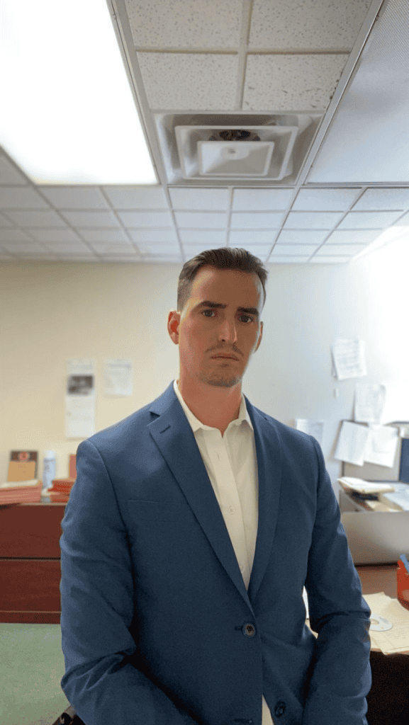 Business professional man in blue suit in office setting with distracted expression| office worker in formal attire looking serious in workplace environment | a man in a blue suit standing in an office with papers and desks around, appearing focused and professional | The image features a young man dressed in a blue suit, standing in an office environment with a distracted or concerned expression. The background includes typical office elements such as desks, papers, and office ceiling tiles, illustrating a professional workspace scenario. | Not relevant.