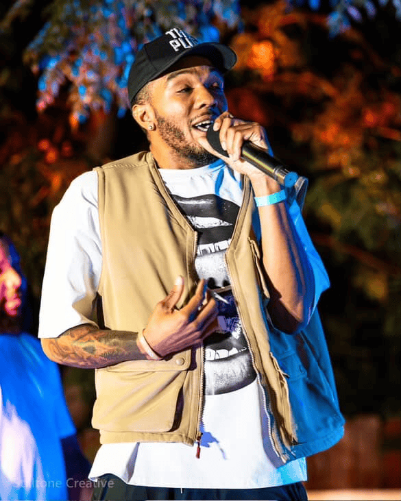 Young male rapper performing on stage at night with colorful background, wearing a beige vest, white t-shirt, and black cap, holding a microphone for a live music performance.