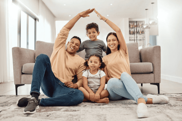 Smiling family of four sitting on the living room floor with their arms raised in a house setting, family fun, togetherness, modern home interior, happy moments, diverse family, cozy living space, indoor lifestyle.