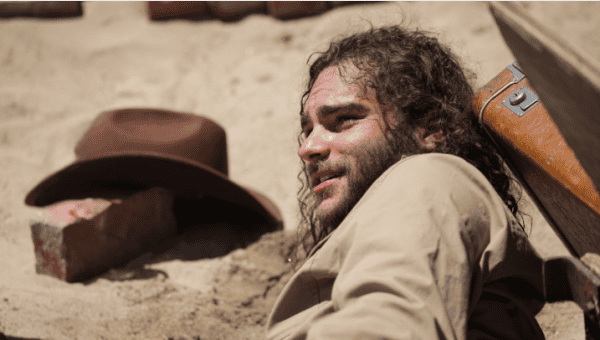 A man with long curly hair and a beard sitting outdoors on a sandy surface, looking relaxed with a brown hat nearby, embodying a rugged, adventurous spirit.