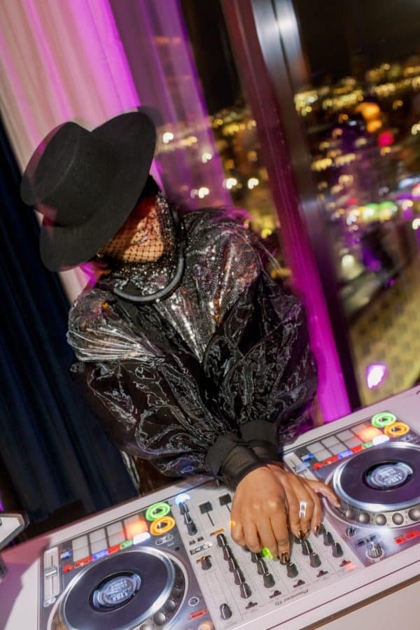 DJ wearing a black hat and shimmering silver outfit mixing music at a nightclub with city lights in the background.